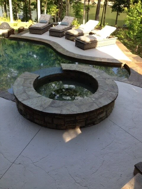 A backyard with a swimming pool and a built-in round fire pit made of stone, surrounded by lounge chairs and trees.