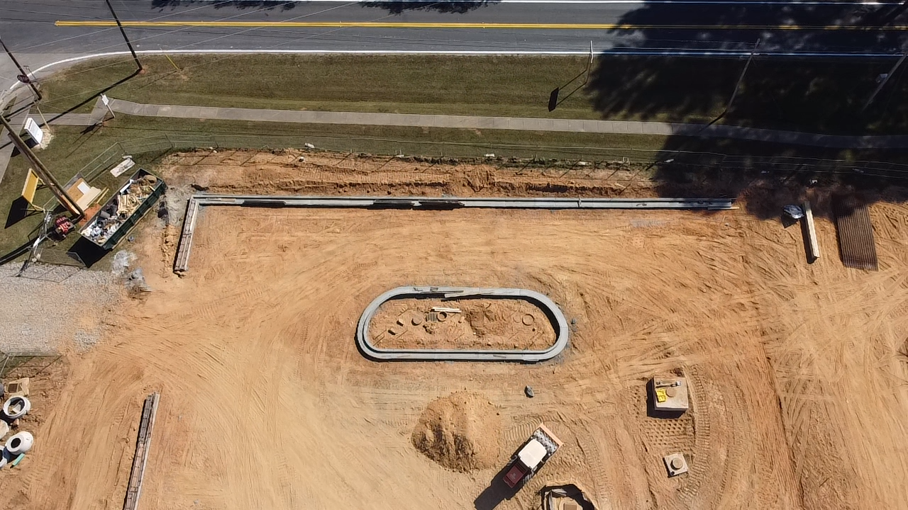 Construction site with a partially built pool outline, construction equipment, and a finished sidewalk next to a road.