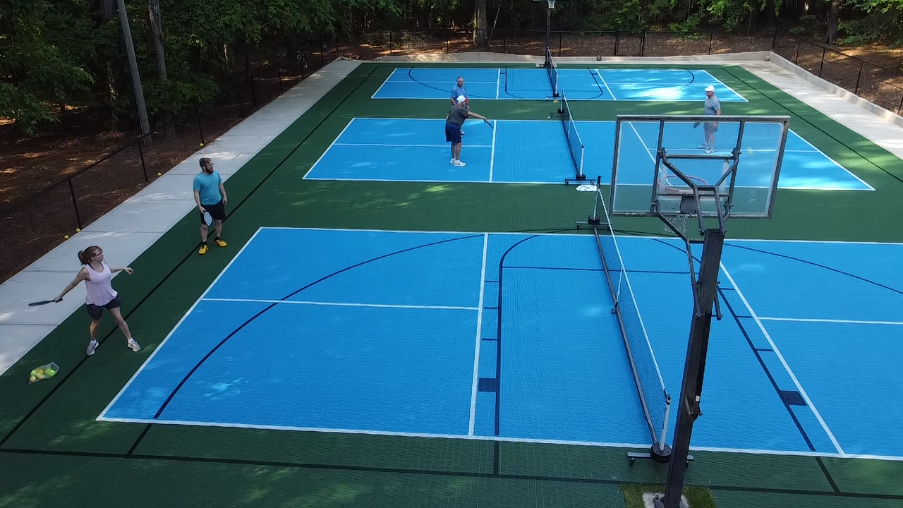 People playing pickleball on an outdoor court with blue playing surfaces and green borders, surrounded by trees.