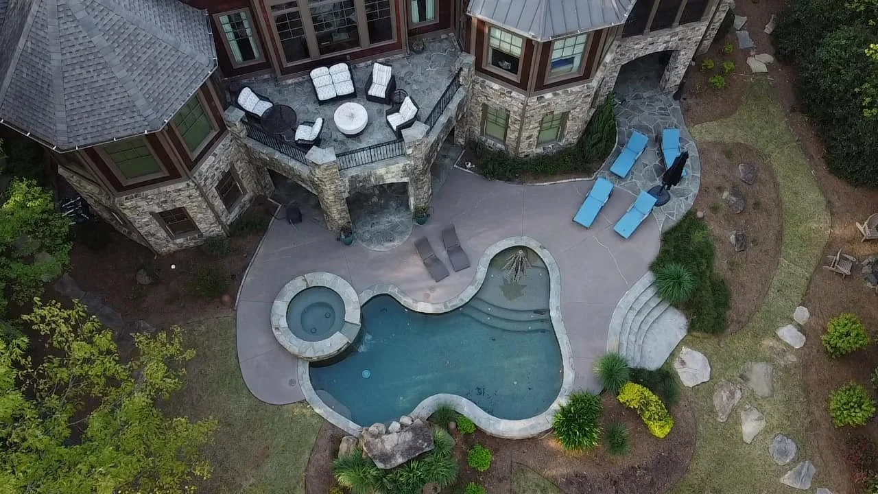 Aerial view of a backyard pool with a hot tub, surrounded by patio furniture, lounge chairs, and landscaped greenery.