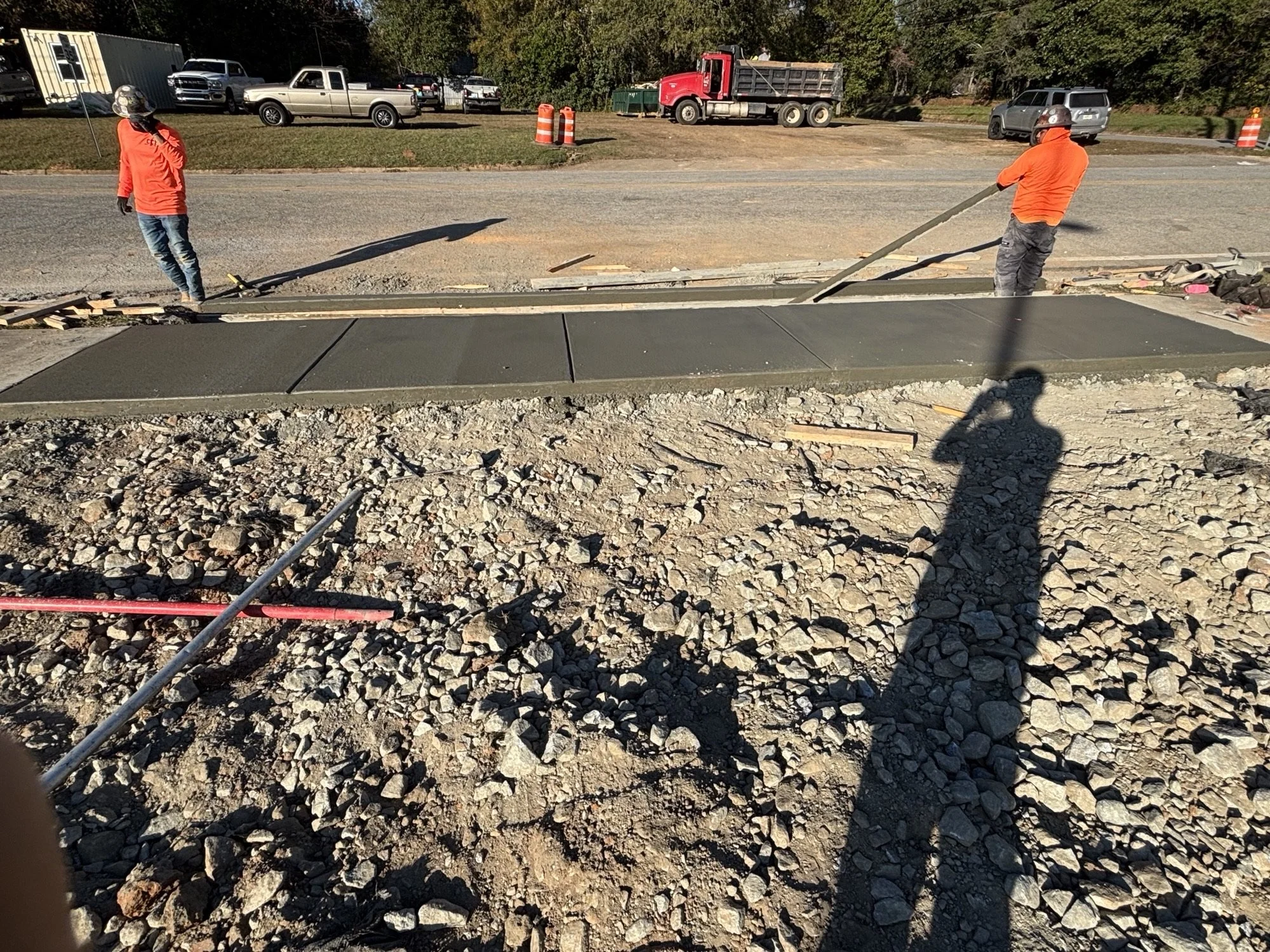 Workers in orange shirts and helmets pouring concrete sidewalk in a construction site, with gravel and construction tools around.