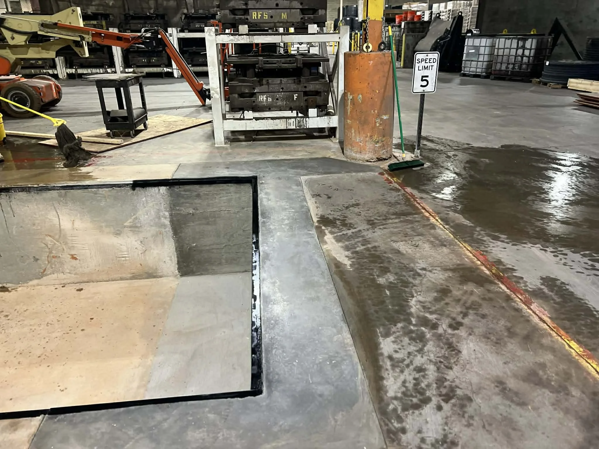 Warehouse or industrial workspace with various equipment, including a cut-out section of floor, a traffic control mirror, a broom, a speed limit sign, storage racks, pallets, and machinery, with wet floor areas.