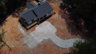 A house with a dark roof and a concrete driveway in front, surrounded by dirt and trees.