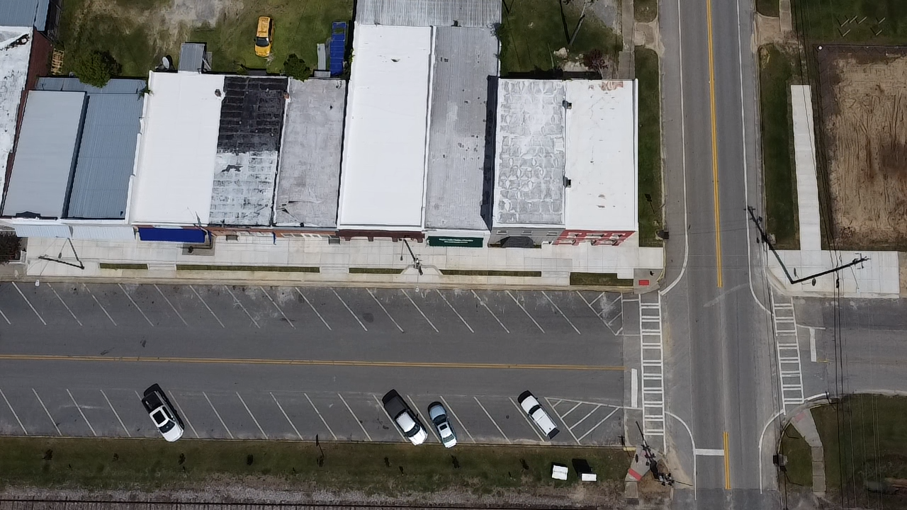 An aerial view of a street with a downtown area featuring several buildings with flat roofs. There is a parking lot with a few parked cars and a crosswalk at an intersection. The surrounding area includes a few trees, grassy patches, and a dirt lot.