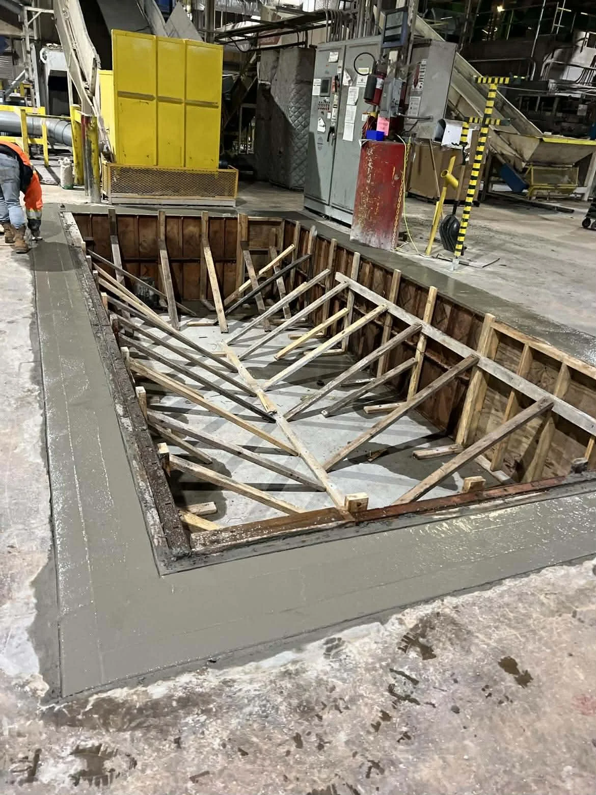 Open excavation site with wooden framing and concrete base, surrounded by industrial equipment and workers in safety gear.