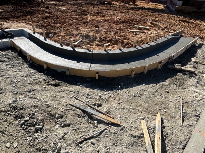 Construction site with a curved concrete foundation for a sidewalk or pathway, surrounded by disturbed soil and wooden formwork.