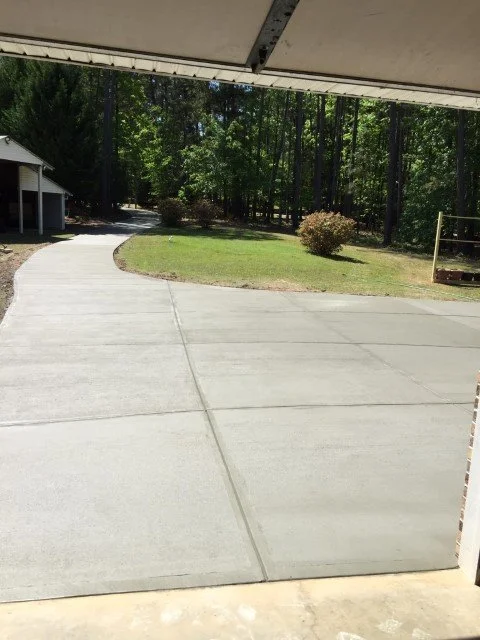 View from inside a garage looking out onto a curved concrete driveway, a grassy area with a small bush, and a wooded yard in the background under a partly open garage door.