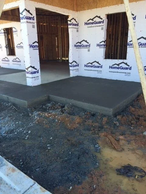 Construction site with a concrete porch in front of a house under construction, with HomeGuard building wrap and framing visible.