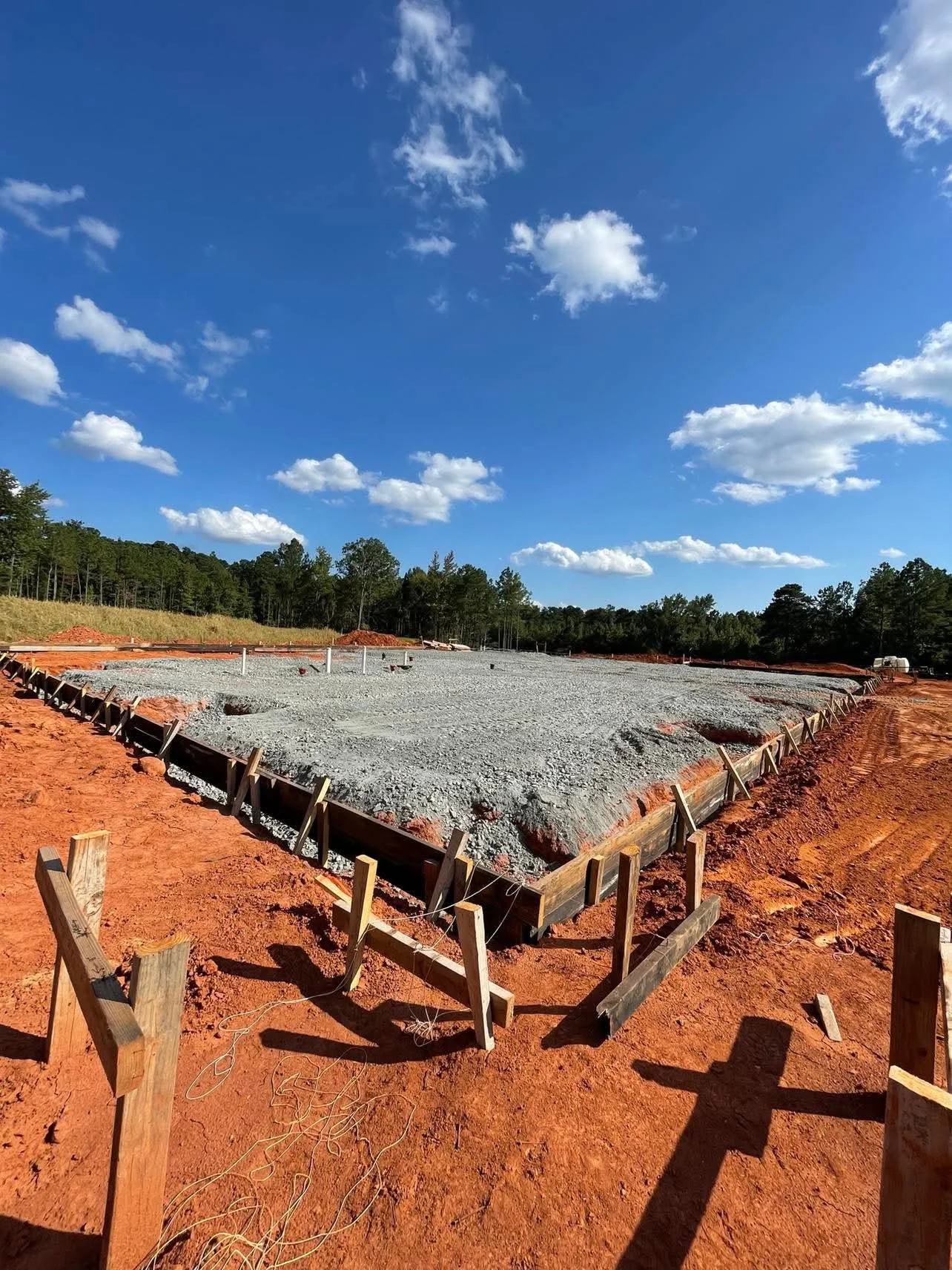 Construction site with a leveled gravel foundation, wooden framing border, and open red soil in foreground, surrounded by trees under a partly cloudy blue sky.