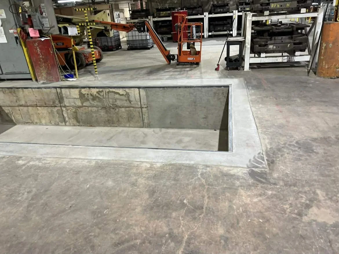 Construction site in an industrial warehouse with a large underground pit, tools, and equipment stored in the background.