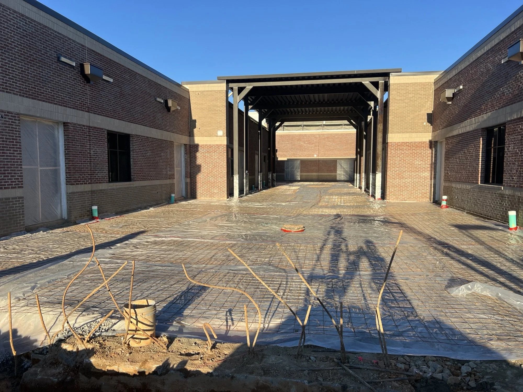Construction site of a building with red and tan brick walls, a covered walkway or entrance with a black metal roof, and exposed rebar on the ground, indicating ongoing foundation work.