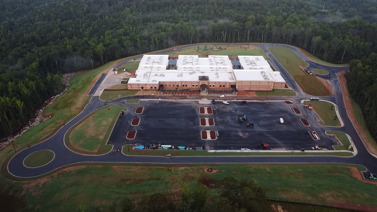 An aerial view of a large, white-roofed building with surrounding roads, parking lot, and greenery, indicating it is under construction or new.