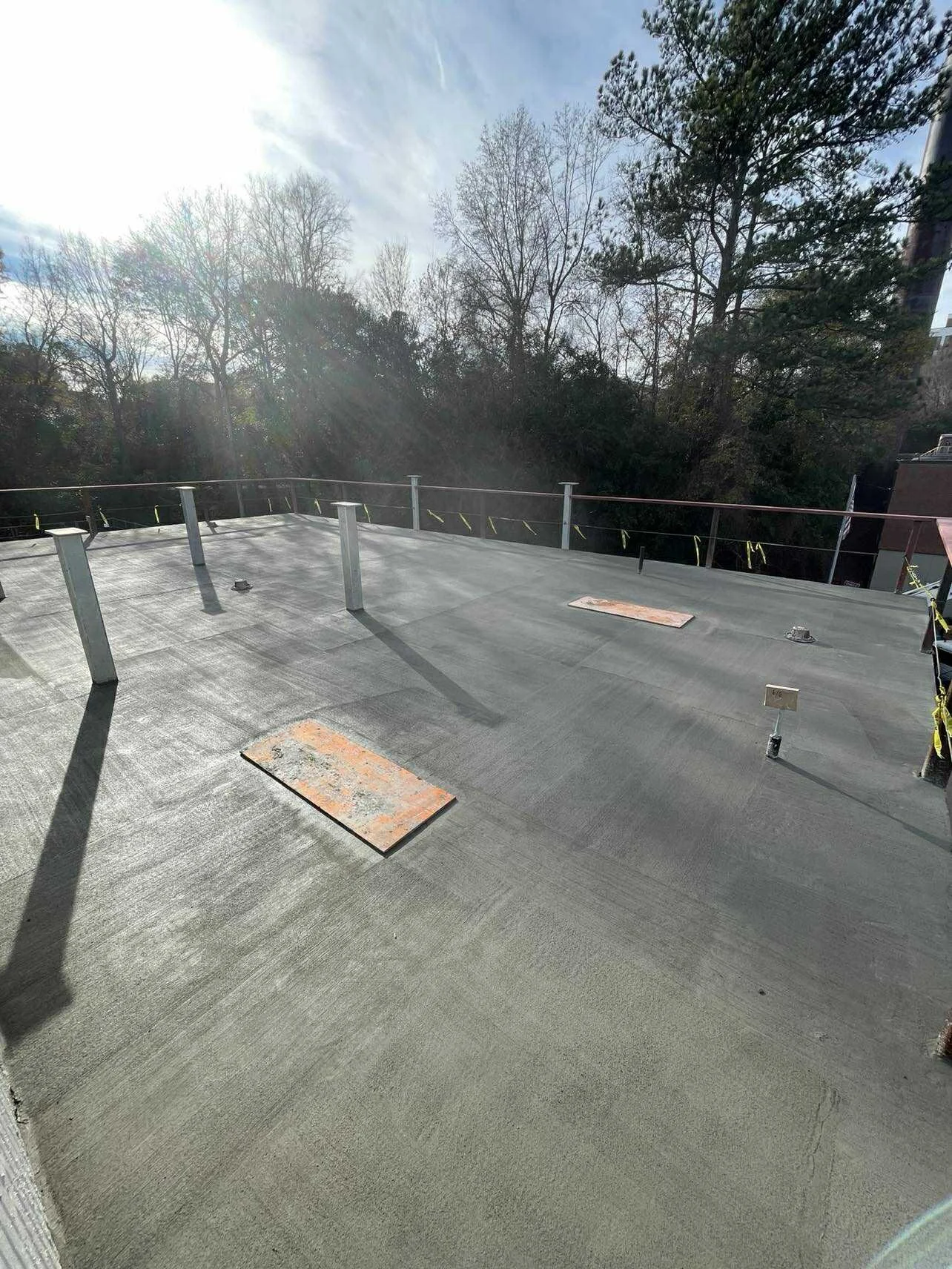 A flat rooftop under construction with metallic vents and access panels, surrounded by trees and a partly cloudy sky.