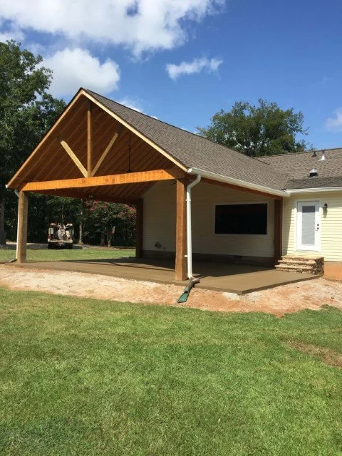 A house with a newly constructed covered patio area with a gabled roof, wood supports, and steps leading up to a door on the side of the house, with a grassy yard and trees in the background.
