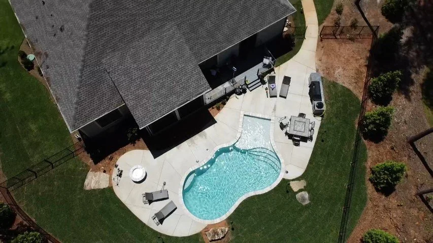 Aerial view of a backyard with a kidney-shaped swimming pool, patio furniture, and a house with a gray roof, surrounded by a lawn and trees.
