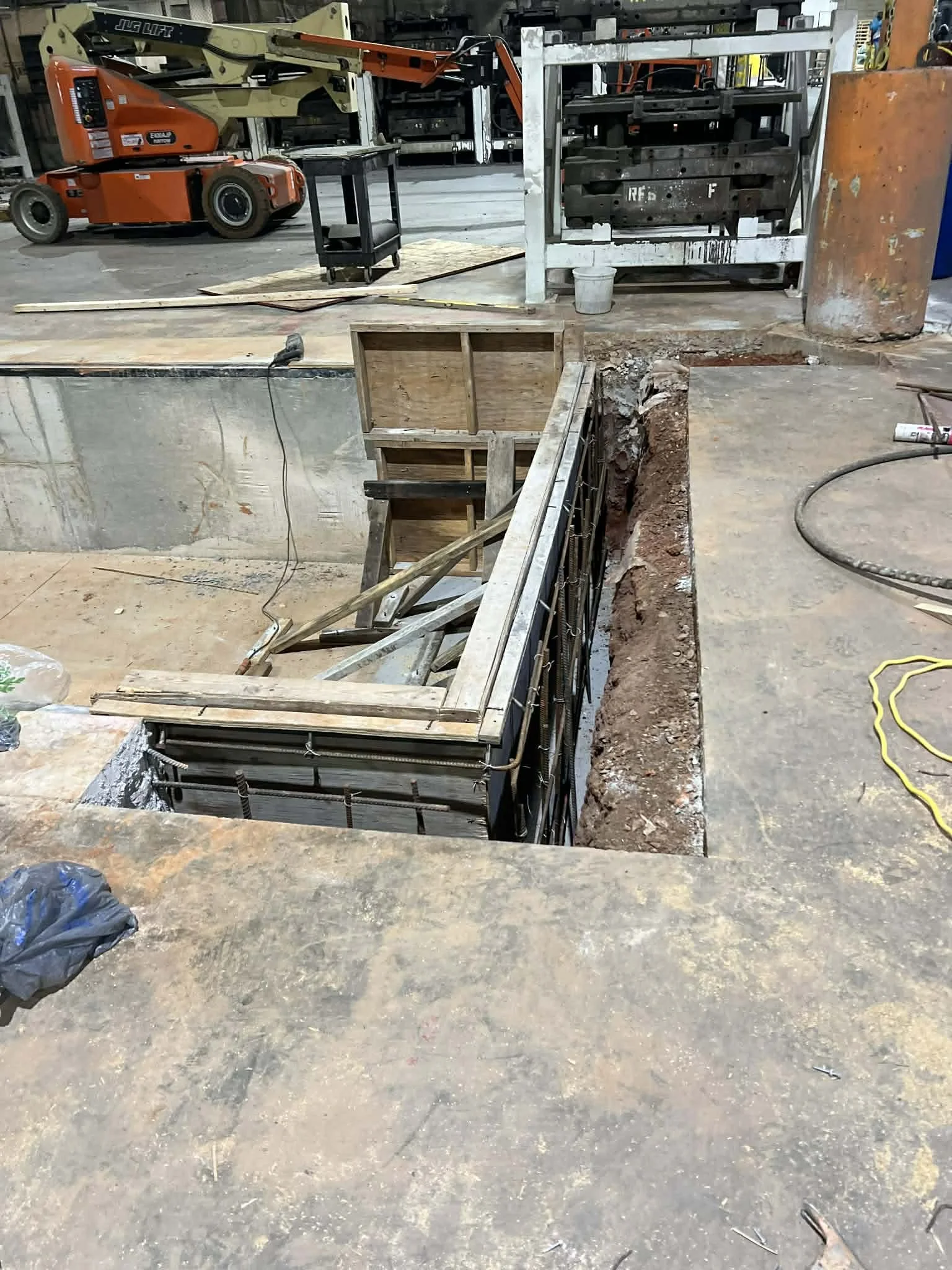 Construction site with a large rectangular hole in the ground, surrounded by steel rebar on one side and a wooden frame on the other. Construction tools and materials are scattered around, including a trash bag, a power drill, and various pieces of l