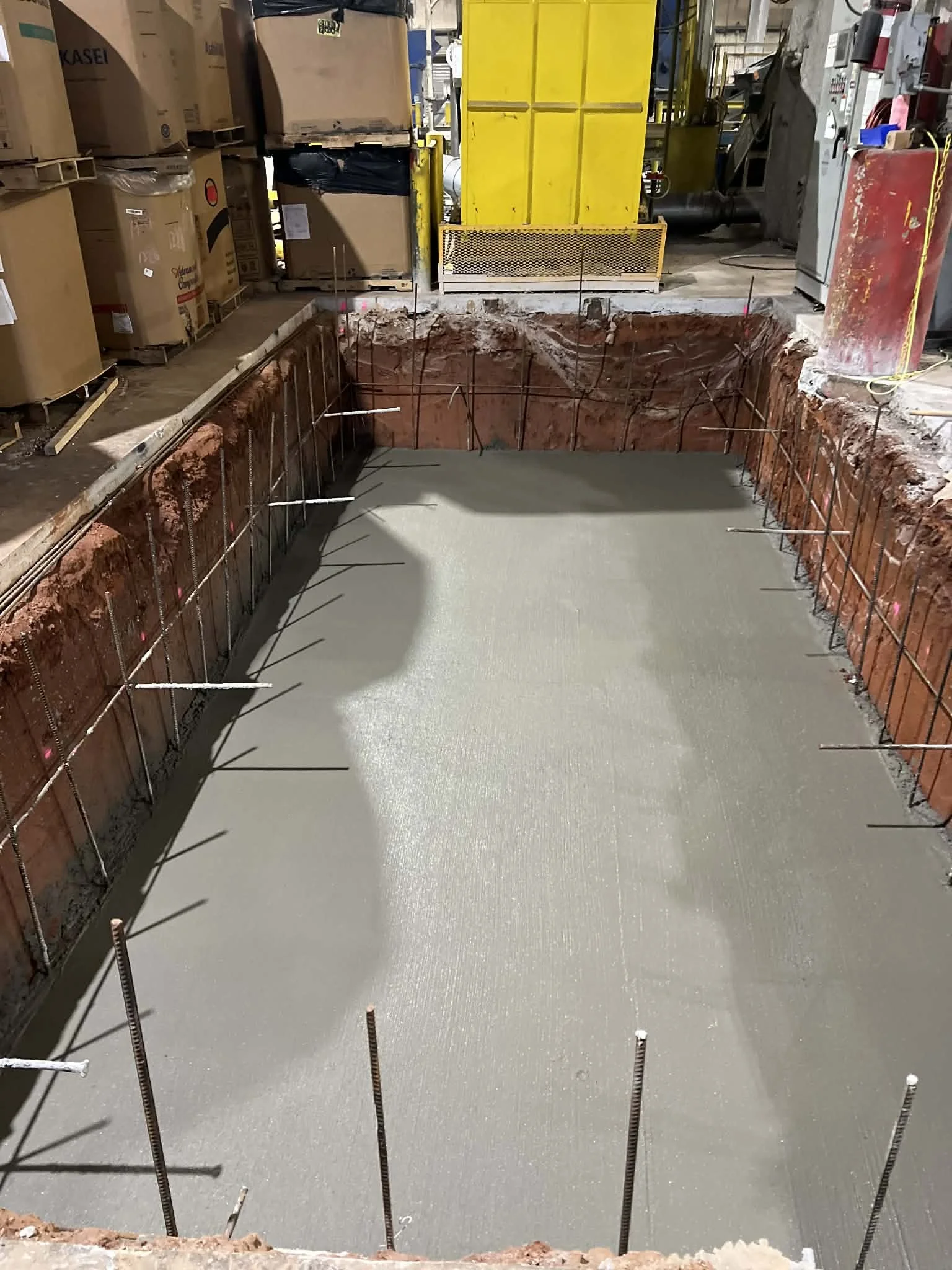 Construction site with freshly poured concrete in a rectangular area, surrounded by rebar, and construction materials and equipment around.