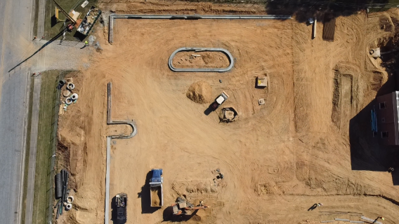Construction site with dirt, equipment, and pipes, and a partially built structure. Boarded-up building on right side.