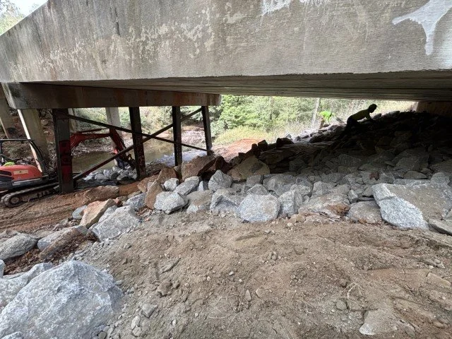 Construction site under a bridge with rocks and dirt, construction equipment, and trees in the background.