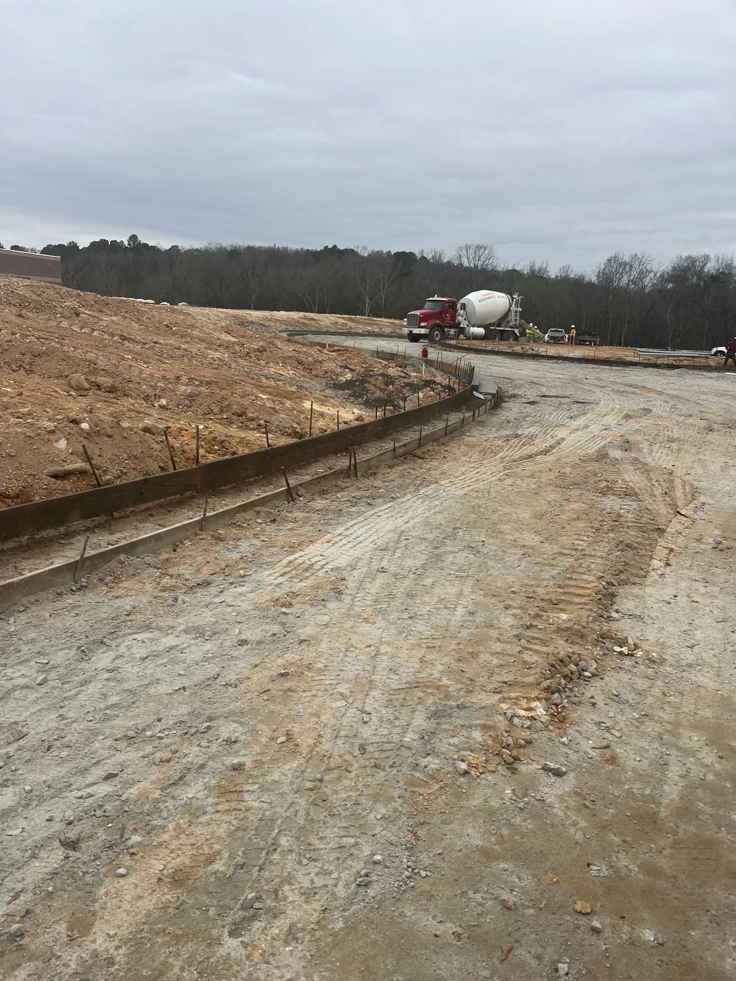Construction site with dirt road, construction workers, a concrete mixer truck, and vehicles. Overcast sky with cloudy weather.