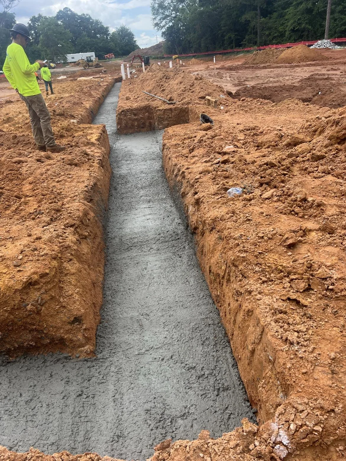Construction workers pouring concrete into a freshly dug trench at a construction site, with equipment and dirt mounds in the background.