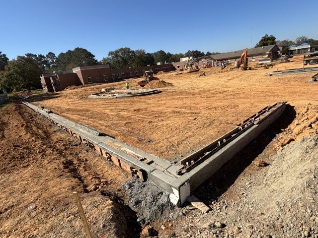 Construction site with a long concrete curb and dirt, residential buildings in the background, and clear blue sky.