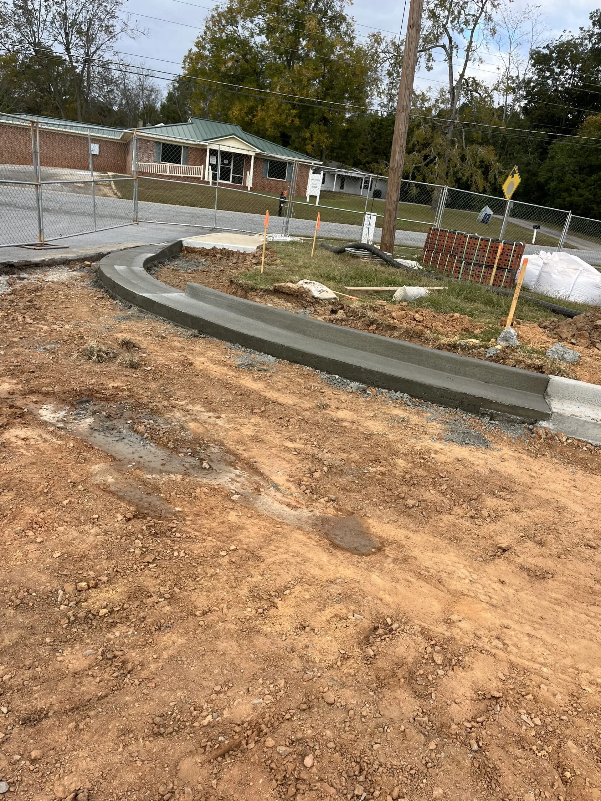 A construction site with a curved section of fresh concrete on the ground, surrounded by dirt. Behind, there is a chain-link fence, a utility pole, stacked bricks, and construction materials. In the background, there are residential houses, a yard wi