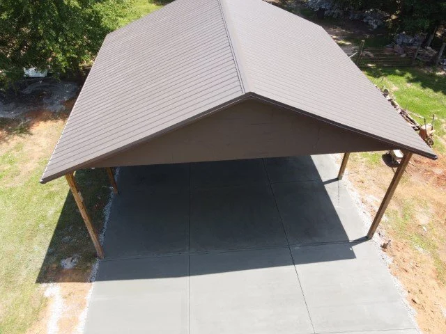 A large, open outdoor pavilion with a metal roof and four wooden posts, situated on a concrete slab in a grassy area with trees nearby.