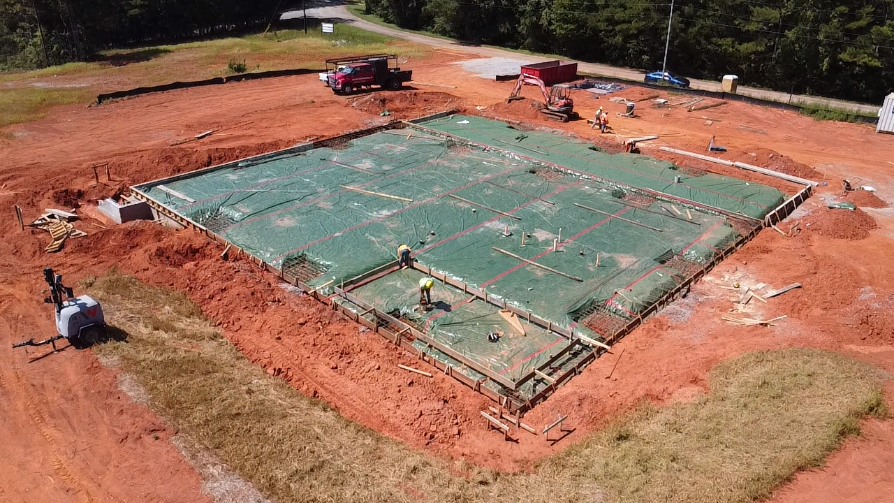 Construction site with a foundation for a building, covered with green plastic sheeting, workers, and construction equipment.