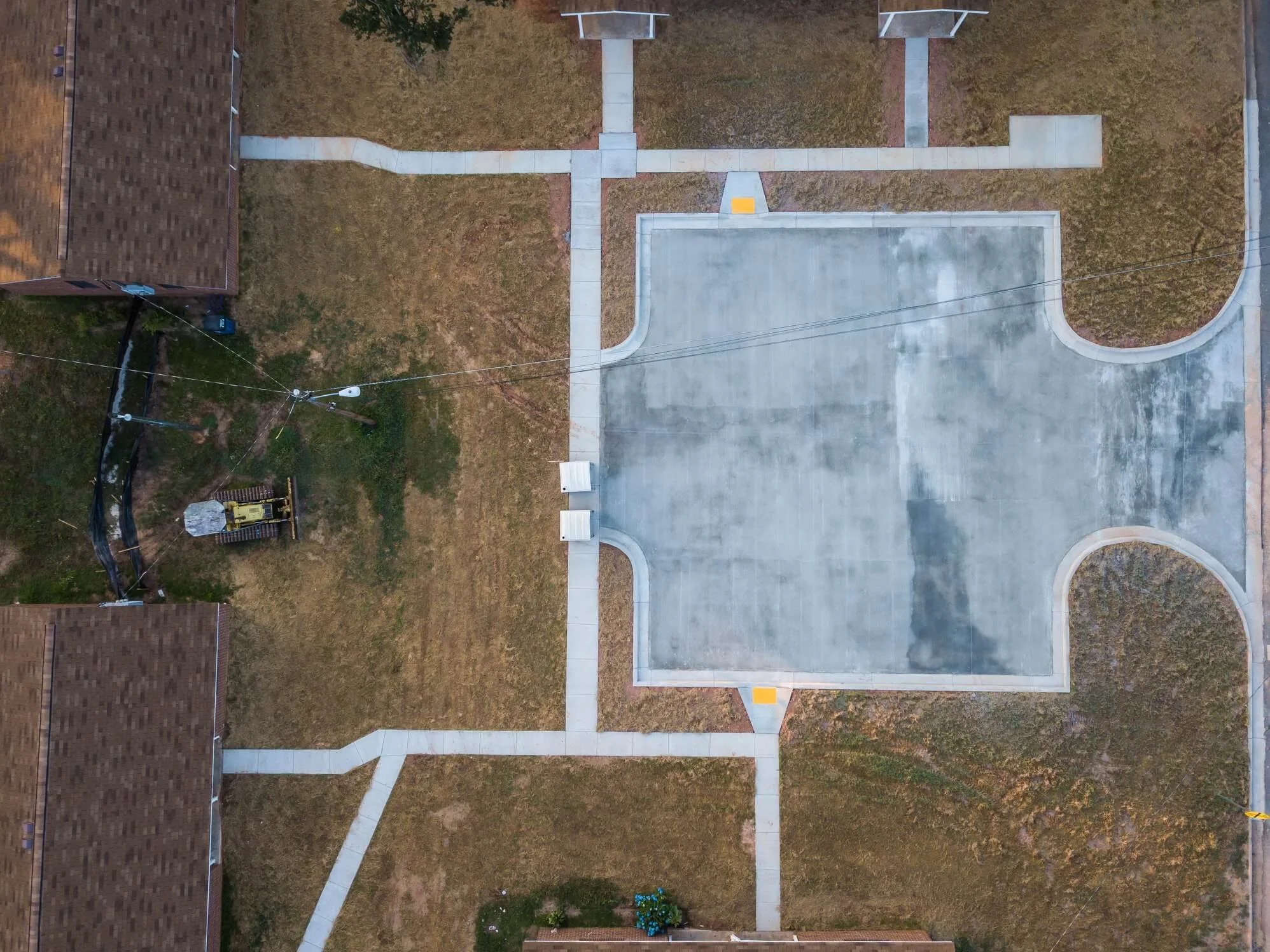 An aerial view of a residential backyard showing a concrete patio with curved borders, a walkway dividing the yard, a grass area, a brick building, a small garden with flowers, a trampoline, a utility box, and utility wires overhead.