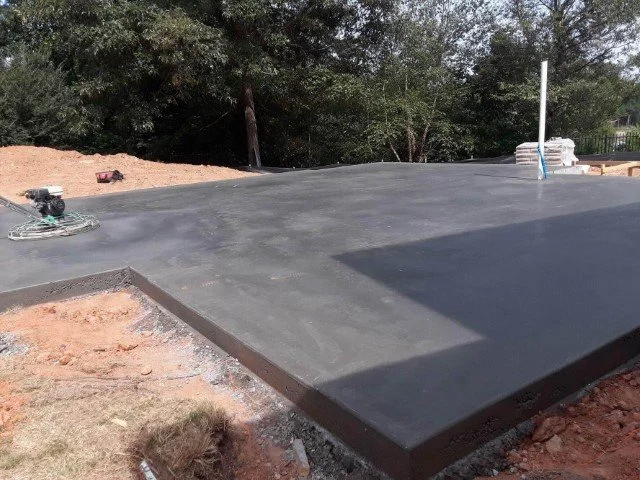 Freshly poured concrete slab on a construction site with power trowel equipment, surrounded by dirt and construction materials.