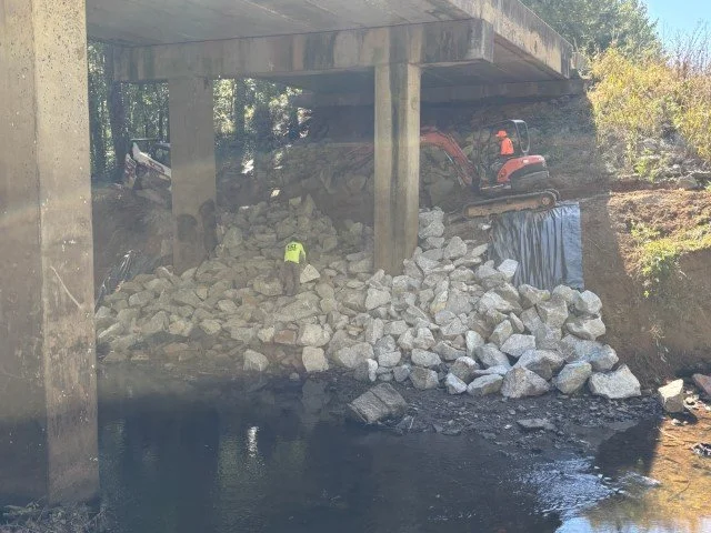 Collapsed rocks and debris under a bridge with a construction worker wearing a yellow vest, and construction equipment nearby.