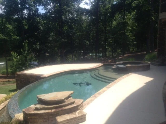 Backyard with a small swimming pool featuring stone accents, surrounded by a concrete patio and lush trees in the background.