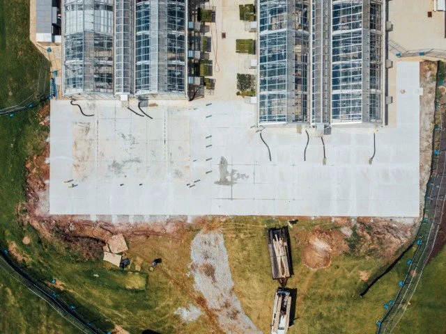 Aerial view of a construction site with a large white concrete slab, nearby green grass, and multiple glass-paneled buildings in progress.