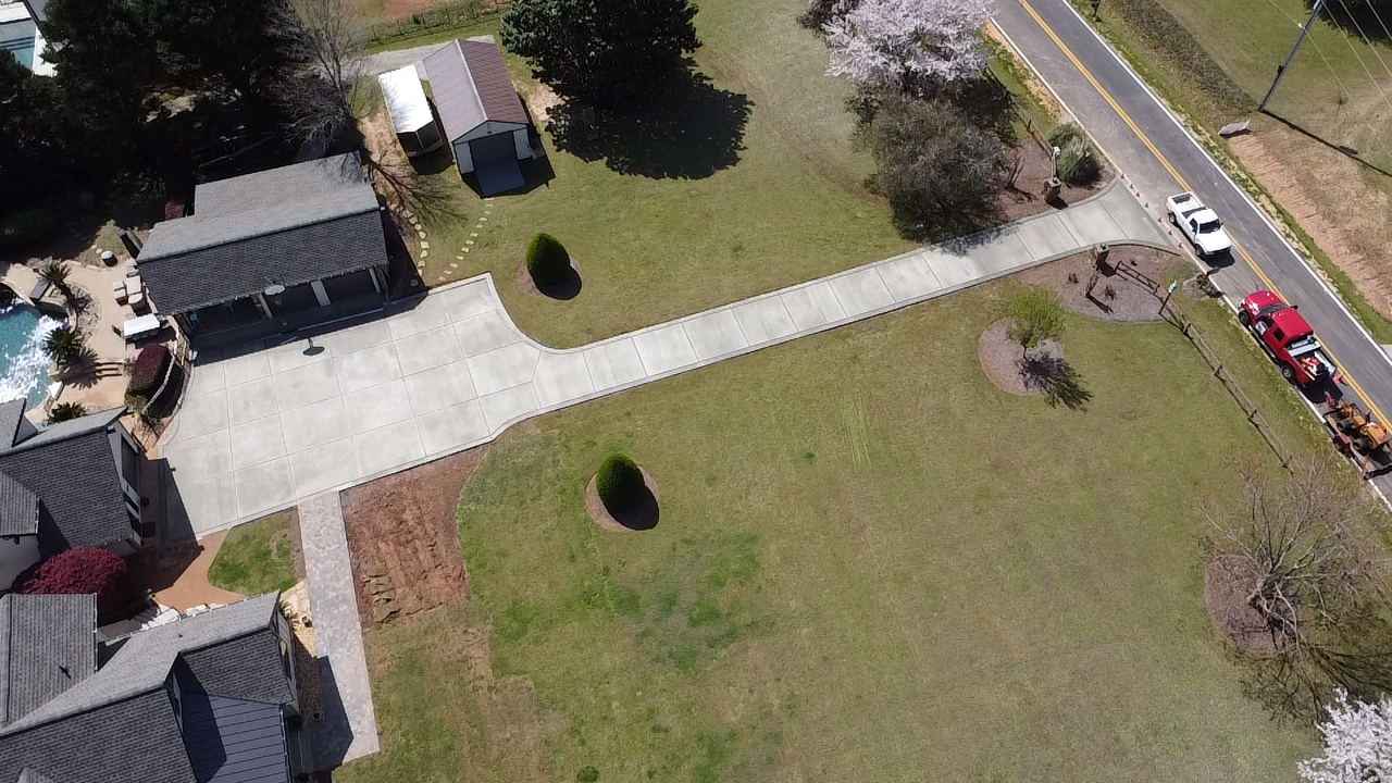 Aerial view of a suburban neighborhood with houses, a driveway, a grassy park area, trees, and parked vehicles along the street.