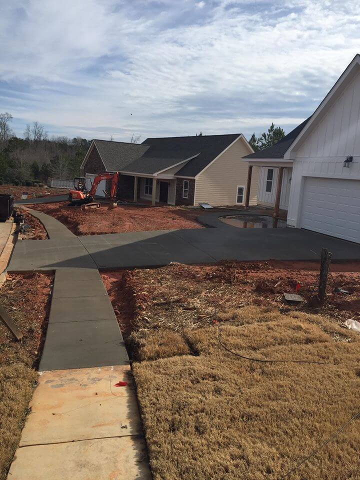 Newly paved driveway leading to a house under construction, with a sidewalk and yard with brown grass and dirt patches, and construction equipment in the background.