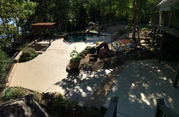 Residential backyard with a hot tub, shaded seating area, garden planters, and a patio with trees casting shadows.