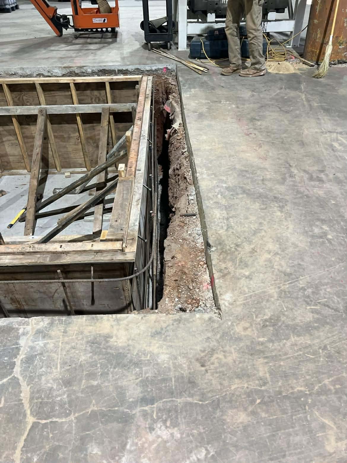 Construction site with an open trench in the floor and unfinished framing for a wall or structure. Workers and construction equipment are visible in the background.