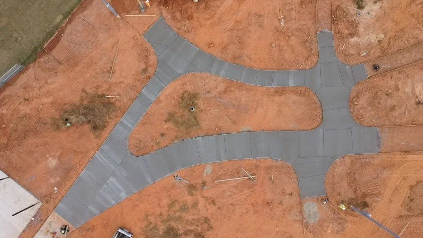 Aerial view of a landscaped area with winding concrete walkways and patches of reddish dirt. No plants or structures are visible, only the walkways and dirt patches.