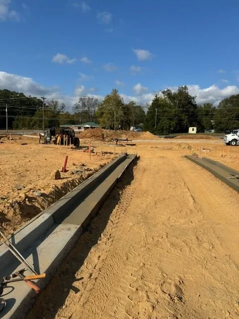 Construction site with concrete curbs being installed, dirt ground, trees in the background, and a blue sky with clouds.