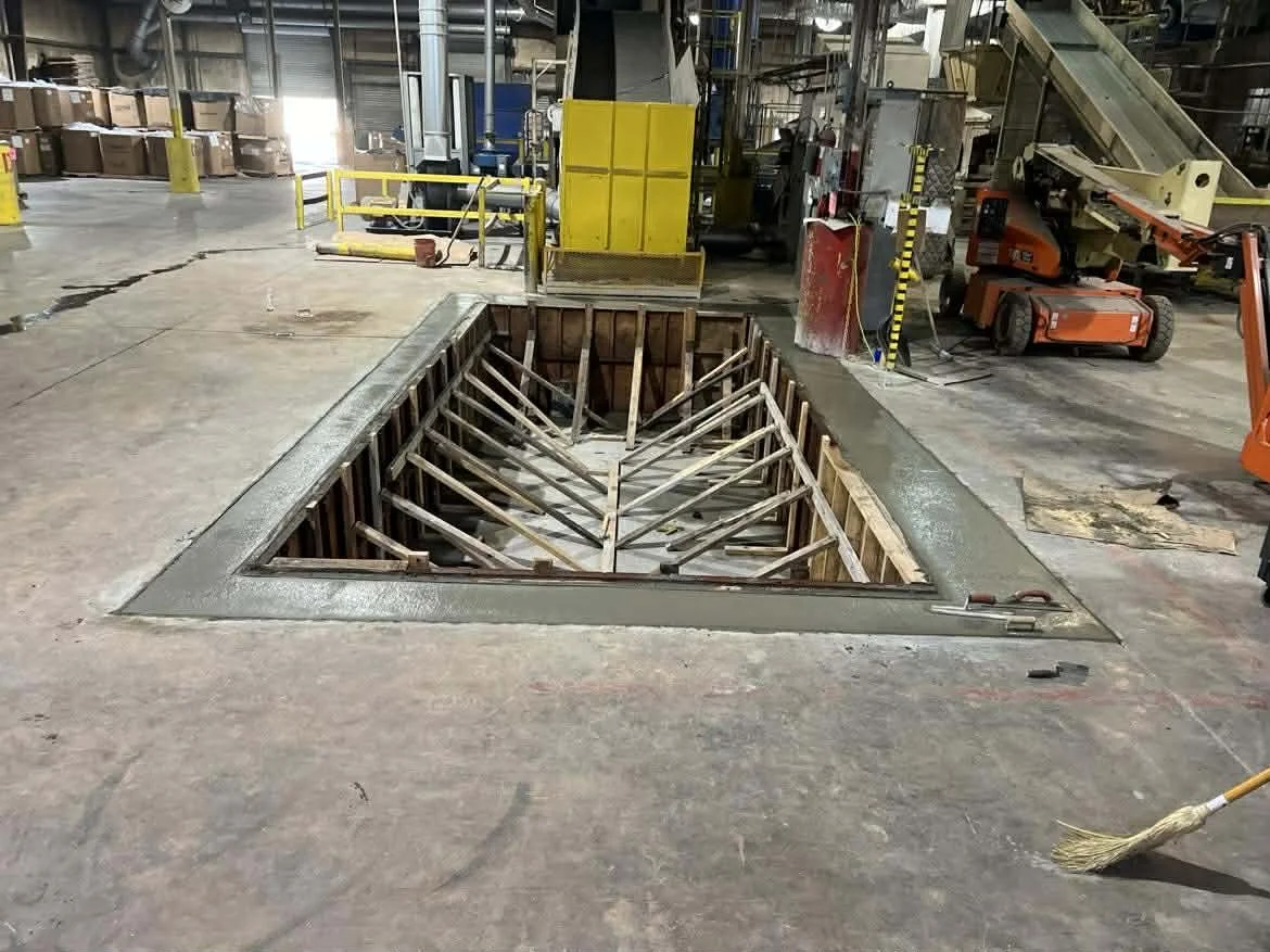 A large square hole in a warehouse or industrial floor, with wooden and metal supports inside, possibly for a staircase or elevator installation. Construction tools and machinery are visible around the area.