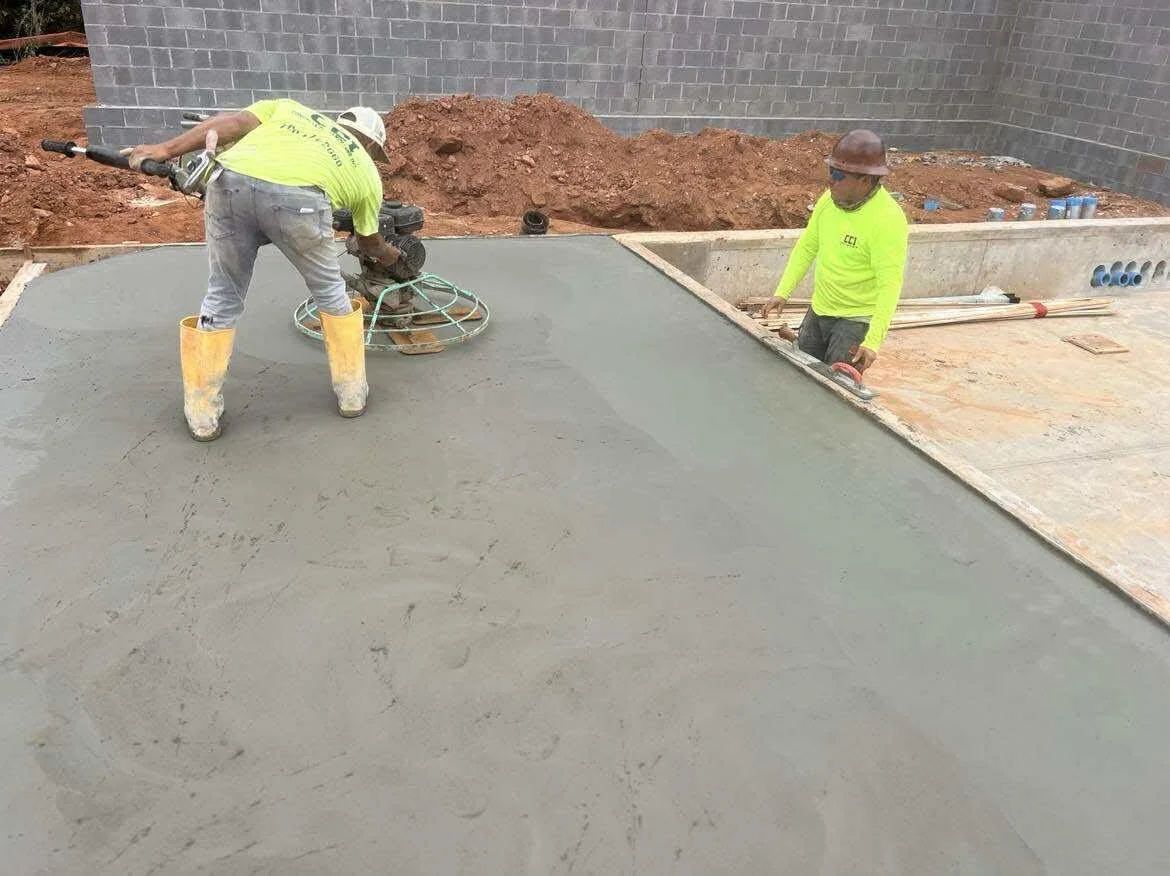 Two construction workers are smoothing freshly poured concrete on a building site, with one using a power trowel and the other observing.