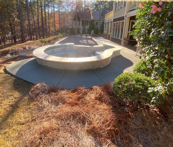 A backyard pool with a kidney-shaped design, surrounded by a concrete deck, adjacent to a house with large windows and a porch. There are trees in the background and shrubs on the right side, with pine straw mulch on the ground.