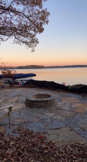 A lakeside scene at sunset with a large tree in the foreground, a stone fire pit, a wooden bench, and calm water reflecting the colorful sky.