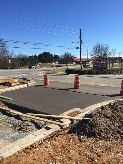 Construction site with freshly poured concrete, two orange traffic barrels, and wooden forms, in a parking lot area near a busy street.