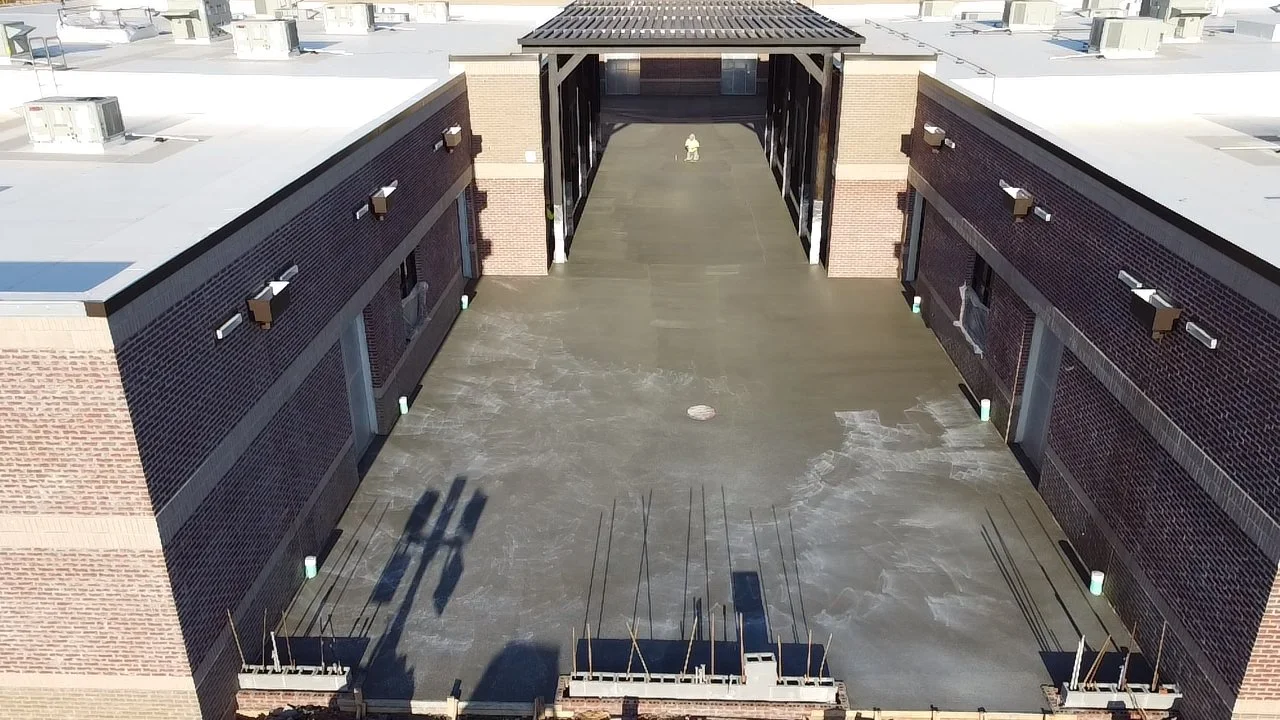 An aerial view of the rooftop of a commercial building under construction, showing the rooftop surface, building walls, and a structure with a roof in the center.