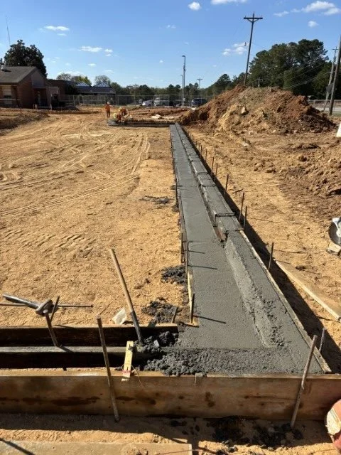 Concrete sidewalk being constructed along a dirt pathway at a construction site with a grassy hill, power lines, and trees in the background.