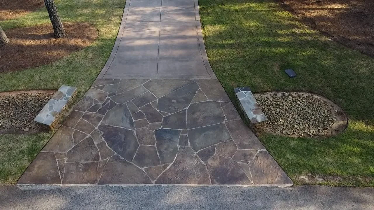 A concrete sidewalk with a stamped stone pattern leading to a lawn and a tree on the left side, with small rock patches on either side.