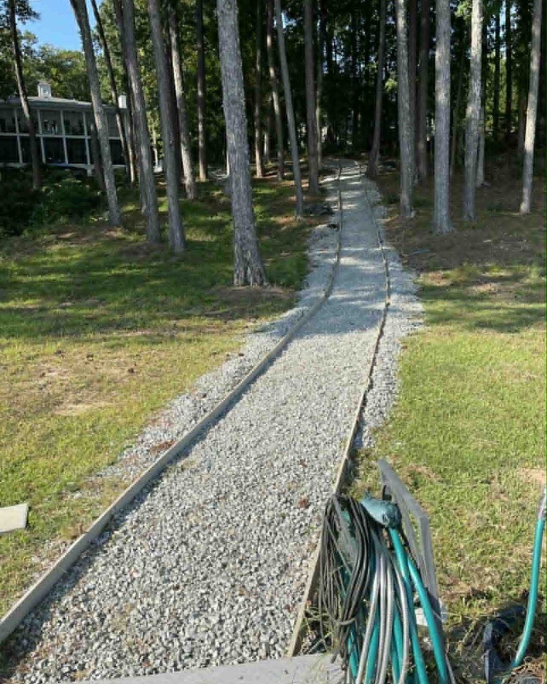 A narrow gravel pathway leading through a wooded area with tall trees and some grass on the sides, with a house visible in the background to the left.