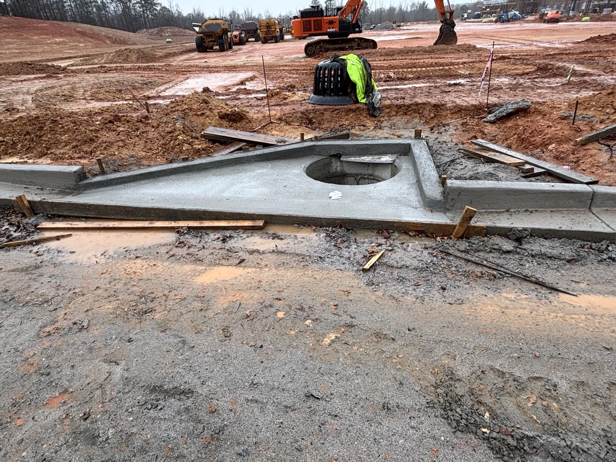 Construction site with excavator, various construction vehicles, and a large concrete structure in the foreground. Soil and dirt are visible everywhere, and construction personnel are present.